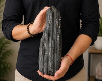 May include: A large, raw black tourmaline crystal specimen with a vertical, striated, and columnar structure. The mineral features a dark, opaque, and jagged surface with natural ridges and a pointed, crystalline top formation.