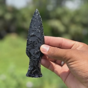以下が含まれることがあります： 手持ちの黒曜石の矢じりで、鋭い三角形の先端が付いています。矢じりは質感のある表面と暗く光沢のある外観をしています。背景はぼやけており、緑の葉が見えます。