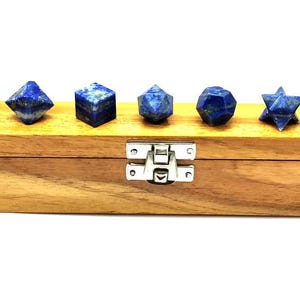 May include: A wooden box with a silver clasp holds seven blue lapis lazuli gemstone shapes. The shapes include a pyramid, a diamond, a cube, a dodecahedron, a icosahedron, a star, and a sphere. The box is made of light brown wood.