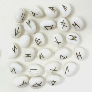 Op de afbeelding: Een verzameling witte, ovale runenstenen met donkere markeringen. Elke steen heeft een ander symbool, waarschijnlijk afkomstig van een oud alfabet of divinatiesysteem. De stenen liggen op een wit oppervlak, wat wijst op een ritueel of presentatie.