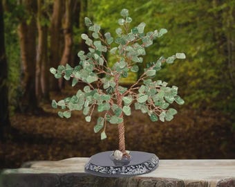 May include: A decorative tree sculpture with a copper wire trunk and branches, adorned with green gemstone leaves. The tree sits on a dark gray, round base. The background is a blurred forest scene.