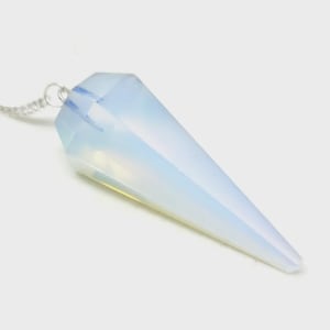 Puede incluir: Un péndulo de cristal facetado y opalescente con una cadena plateada. El cristal es de color pálido y translúcido, con forma cónica y puntiaguda. La cadena es delicada y está unida a la parte superior.