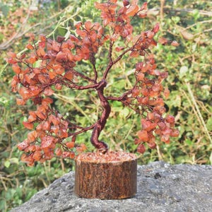 May include: A decorative wire tree sculpture with orange gemstone leaves. The trunk is twisted copper wire, and the base is a wooden cylinder. The tree sits on a grey rock, with a blurred green backdrop.