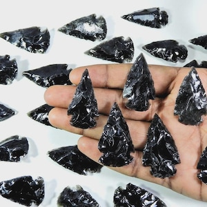 Puede incluir: Colección de puntas de flecha de obsidiana negra, algunas sostenidas en una mano, otras dispersas sobre una superficie blanca. Las puntas de flecha tienen una superficie brillante y reflectante y están intrincadamente formadas con puntas afiladas y bordes detallados.
