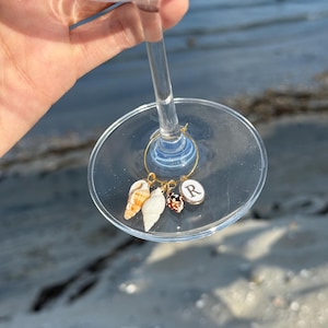 May include: A clear wine glass with a gold-coloured charm featuring three seashells and a circular charm with the letter "R". The seashells are various colours and shapes. The glass is held up against a blurred background of sand and water.