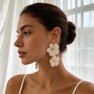 May include: A pair of off-white drop earrings featuring two stacked flower shapes. Each flower is constructed from coiled cord or thread, with a small gold-toned bead at the center of each bloom. The earrings hang vertically from the earlobe.