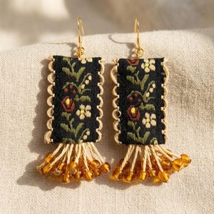 May include: A pair of rectangular drop earrings featuring black fabric with a floral embroidery pattern in red, green, and cream. The edges are trimmed with beige scalloped cord, and the base has beige fringe with small amber-colored beads.