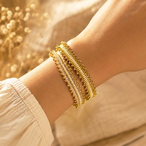May include: Two layered bracelets featuring a central band of white seed beads bordered by rows of small, metallic gold-toned beads. The design incorporates a delicate, fringed edge effect created by the arrangement of the gold beads along the sides.