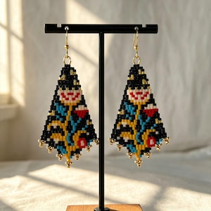 May include: A pair of triangular beaded earrings featuring a geometric floral pattern in black, white, red, yellow, and blue glass beads. The earrings hang from gold-toned metal hooks and are finished with small gold-toned beads along the bottom edge.