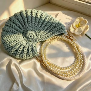 May include: A sage green crocheted handbag with a shell-like spiral texture. The bag features a detachable strap made of white faux pearls and a braided white cord, connected by gold-toned metal clasps. A small white crocheted flower is attached.