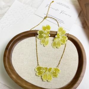 Op de afbeelding: Een kralen sieradenset met een ketting en bijpassende oorbellen. De set heeft een bloemenontwerp in tinten geel en wit, met een klein parelaccent. De ketting en oorbellen zijn aan een gouden ketting bevestigd. Op de achtergrond is een kaart met de tekst "Favourite Flowers" te zien.