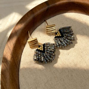 May include: A pair of stud earrings featuring a gold-toned square top with horizontal ridges, connected to a fan-shaped fringe of small, cylindrical beads in shades of metallic grey, black, and gold, arranged in a layered, radiating pattern.