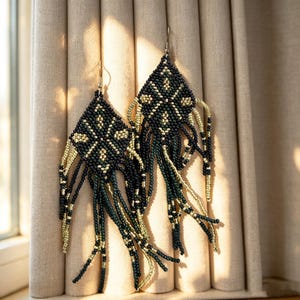 May include: A pair of diamond-shaped dangle earrings crafted from small black and metallic gold seed beads. The earrings feature a geometric pattern in the center and long, flowing beaded fringe strands hanging from the bottom edges.