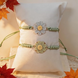 May include: Two handmade crochet bracelets featuring sage green braided bands and central flower motifs. The top bracelet has a light gray flower with a yellow center, while the bottom bracelet features a sage green flower with a yellow center.