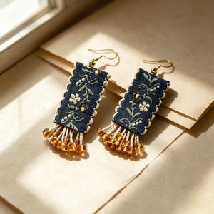 May include: A pair of rectangular drop earrings featuring dark blue fabric with an embroidered floral pattern in white, green, and yellow. The edges are finished with cream-colored trim, and the bottom features dangling amber-colored glass beads.