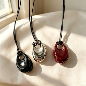 Puede incluir: Tres colgantes en forma de lágrima que cuelgan de collares de cordón negro. Un colgante es negro brillante, otro es de metal plateado pulido y el tercero es de color rojo oscuro. Cada colgante tiene una abertura circular en la parte superior.