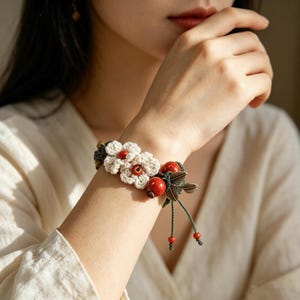 May include: A bracelet featuring two cream-colored crocheted flowers with small red beads at their centers, accented by larger red spherical beads and metallic bronze-toned leaf charms, all strung on a dark green cord with adjustable dangling ends.