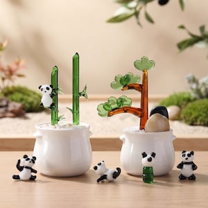 Peut inclure: Deux pots en céramique blanche contenant des sculptures de plantes en verre. Un pot contient des tiges de bambou en verre vert avec une petite figurine de panda, tandis que l'autre présente un arbre en verre brun et vert. Quatre pandas sont posés.