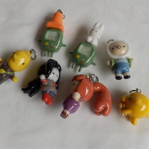 Può includere: Una collezione di sette piccoli ciondoli a forma di personaggio in argilla, lucidi e multicolori. I ciondoli raffigurano varie figure, tra cui una console verde, un coniglio bianco, un personaggio con cappuccio bianco e altre forme con anelli metallici.