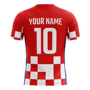 Puede incluir: Camiseta de f&uacute;tbol roja y blanca con el texto "YOUR NAME" sobre el n&uacute;mero "10" en blanco. La camiseta tiene un patr&oacute;n de cuadros en la mitad inferior y las mangas. Es una camiseta de manga corta.