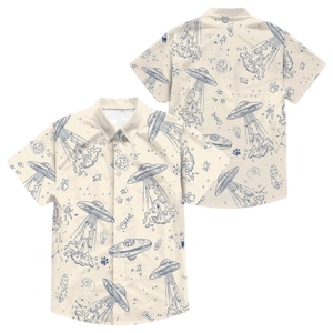 May include: A short-sleeved button-down shirt with a cream-colored background and a repeating pattern of blue UFOs, planets, stars, and other space-themed doodles. The shirt has a classic collar and button closure.