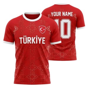 Op de afbeelding: Rood voetbalshirt met witte randen en het woord "TÜRKIYE" in witte blokletters. Het shirt heeft een Turks vlagembleem en een herhalend geometrisch patroon. De achterkant toont nummer 10 en de tekst "YOUR NAME".