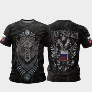 Puede incluir: Camiseta negra con un diseño detallado de oso plateado en la parte delantera y la palabra "RUSSIA" con un escudo de armas en la espalda. La camiseta tiene un patrón de camuflaje y una pequeña bandera en la manga.