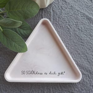 Könnte beinhalten: Ein hellrosa dreieckiges Tablett mit dem Text "SO SCHÖN, dass es dich gibt" in schwarzer Schrift. Das Tablett hat ein marmorähnliches Muster. Eine weiße Vase und grüne Blätter sind im Hintergrund. Das Tablett liegt auf einer grauen Oberfläche.