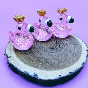 May include: Three pink glass flamingo figurines with gold crowns and black and white beaks sit on a wooden slice. The flamingos have glitter inside, and the background is a solid purple color.