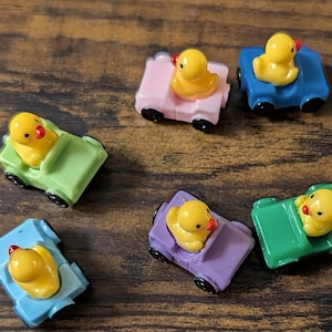 May include: A collection of colorful toy cars, each with a yellow duck figurine as a passenger. The cars are in various colors, including pink, blue, green, and purple. The ducks have red beaks and are sitting in the cars.