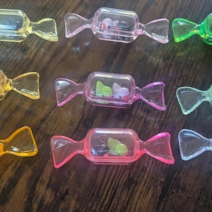May include: A collection of colorful, candy-shaped containers. Each clear container holds small decorative elements, with colors including yellow, pink, green, and clear. The containers are arranged on a dark wooden surface.