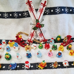 May include: A collection of miniature Christmas figurines, including snowmen, ducks, and reindeer, arranged on a white towel with a snowflake border. Two decorative skis with red snowflake designs are crossed at the top. A festive holiday theme.