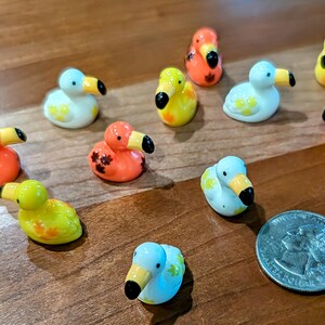 May include: A collection of small, colorful duck figurines in various colors, including orange, yellow, white, and blue. Each duck has a black beak and is decorated with floral patterns. A quarter is included for size comparison.
