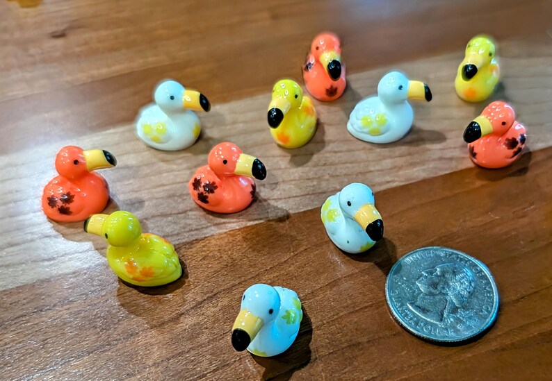 May include: A collection of small, colorful duck figurines in various colors, including orange, yellow, white, and blue. Each duck has a black beak and is decorated with floral patterns. A quarter is included for size comparison.