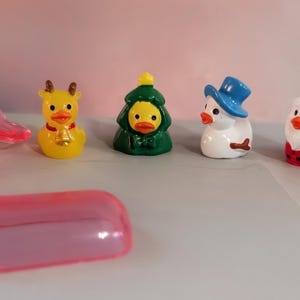 May include: A collection of Christmas-themed rubber duck figurines. The ducks are dressed as a reindeer, Christmas tree, snowman, Santa Claus, and a duck with a top hat. A pink candy wrapper and a pink capsule are in the foreground.