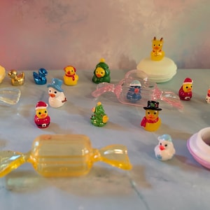 May include: A collection of miniature Christmas-themed figurines, including ducks, snowmen, and penguins, some wearing Santa hats. The figurines are arranged on a light blue surface, with a clear candy-shaped container in the foreground.