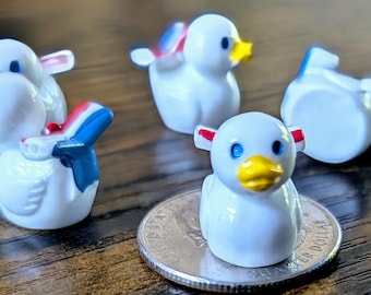 May include: A collection of miniature white duck figurines with blue eyes and yellow beaks. Some ducks have red, white, and blue accents on their wings. One duck is perched on a coin. The ducks are on a wooden surface.