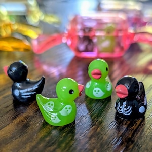May include: A collection of small, colorful duck figurines with Halloween-themed designs. The ducks are in various colors, including black, green, and purple, with white skeleton and ghost patterns. Some are in clear plastic containers.