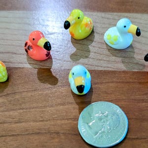 May include: A collection of small, colorful duck figurines with black beaks and various patterns. The ducks are in shades of yellow, orange, and blue. A US quarter is included for size comparison. The figurines are on a wooden surface.