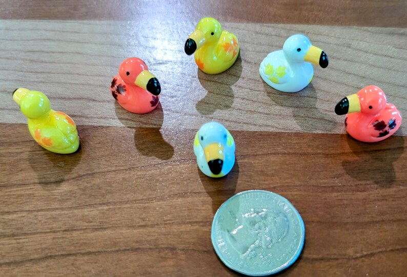 May include: A collection of small, colorful duck figurines with black beaks and various patterns. The ducks are in shades of yellow, orange, and blue. A US quarter is included for size comparison. The figurines are on a wooden surface.