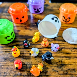 May include: A collection of miniature plastic Halloween-themed items including three small pumpkin-shaped buckets in orange and purple, a white ghost-shaped bucket, and several tiny rubber ducks featuring various colors and festive costumes.