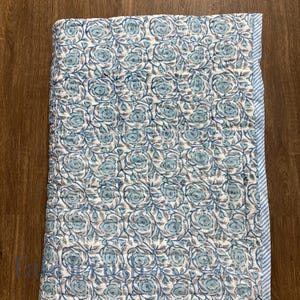 Top view of the Blue Rose Meadow Hand Block Printed Quilt laid flat, showcasing repeating blue rose and floral vine motifs hand-block printed on a soft ivory cotton base. The image highlights detailed quilting, uniform pattern alignment, and a neatly finished blue and white striped border along the edge, emphasizing handcrafted craftsmanship, reversible design, and elegant, timeless bedding style.