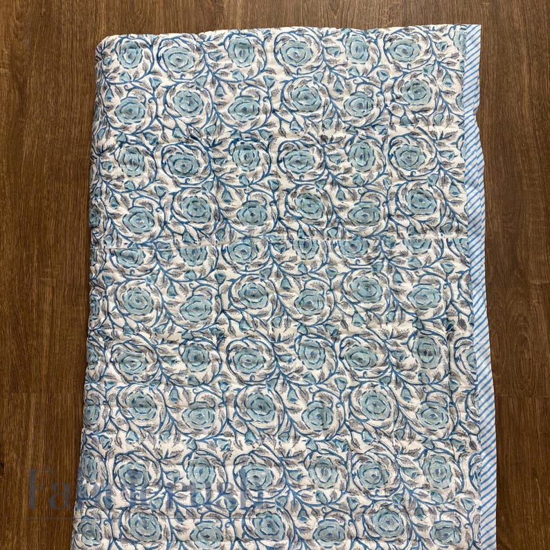 Top view of the Blue Rose Meadow Hand Block Printed Quilt laid flat, showcasing repeating blue rose and floral vine motifs hand-block printed on a soft ivory cotton base. The image highlights detailed quilting, uniform pattern alignment, and a neatly finished blue and white striped border along the edge, emphasizing handcrafted craftsmanship, reversible design, and elegant, timeless bedding style.