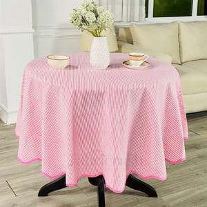 Puede incluir: Un mantel redondo a rayas rosas y blancas con un borde festoneado, que cubre una mesa de madera oscura. Un cuenco pequeño, un jarrón con flores y una taza y platillo están sobre la mesa. El mantel es de tela.
