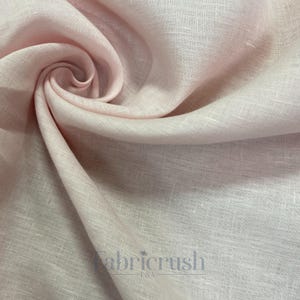 May include: Close-up of a soft, light pink linen fabric. The fabric is tightly woven with a subtle texture and is artfully draped, creating folds and shadows. The Fabricrush USA logo is visible in the lower right corner.