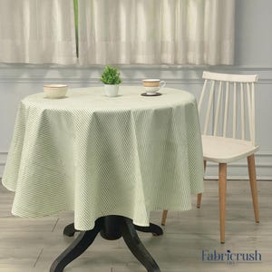 Puede incluir: Un mantel redondo con un estampado de rayas verdes y blancas cubre una mesa oscura. Una silla blanca con patas de madera está al lado de la mesa. Una pequeña planta, un cuenco y una taza de té están sobre la mesa. El logotipo de Fabricrush USA está en la esquina inferior derecha.