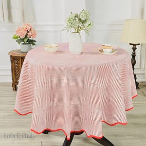 Puede incluir: Un mantel redondo con un estampado de rayas rojas y blancas. El mantel está drapeado sobre una mesa de madera oscura. Un borde rojo bordea el borde del mantel. La mesa está decorada con un jarrón de flores, una taza de té y un cuenco pequeño.