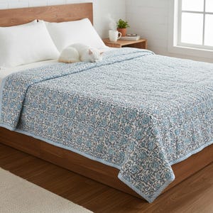 Blue Rose Meadow hand block printed quilt displayed on a wooden bed in a bright, minimal bedroom. The quilt features an intricate blue floral rose pattern on a soft ivory background with a striped border, showcasing traditional artisan craftsmanship. Lightly quilted for comfort, it is neatly spread with a folded edge, creating a cozy, elegant look. A white cat rests peacefully on the quilt, enhancing the warm, homey aesthetic.