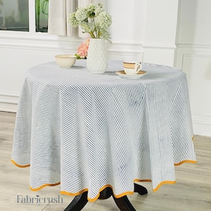 Puede incluir: Un mantel redondo con un estampado de rayas azules y blancas. El mantel tiene un borde amarillo y está sobre una mesa de madera oscura. Un jarrón blanco con flores, un cuenco y una taza de té están sobre la mesa.
