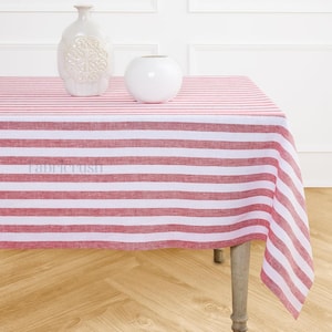 Puede incluir: Un mantel a rayas rojas y blancas cubre una mesa, con dos jarrones de cerámica blanca encima. El mantel tiene rayas horizontales y el texto "fabricrush" es visible. La mesa tiene patas de madera de color claro.